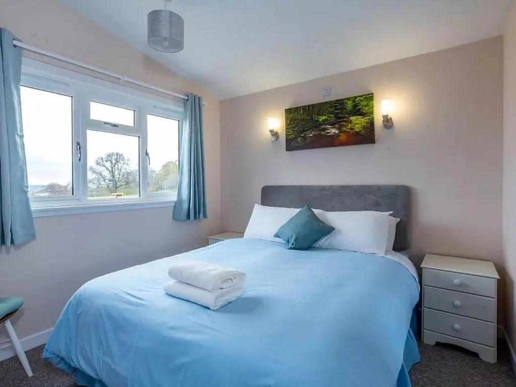 a bedroom with a bed with blue sheets and two windows at Kilninver Chalet on Reelig Estate in Kirkhill