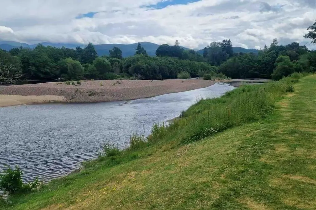 a river with a grassy field next to a field at Coorie Nook, Chalet 103 in Aviemore
