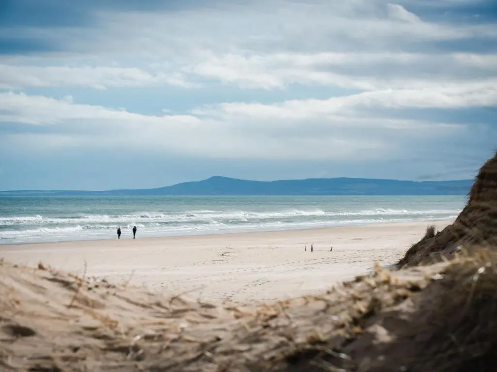 two people walking on a beach near the ocean at Apartment Harbour Nights Deluxe Number 12 by Interhome in Lossiemouth
