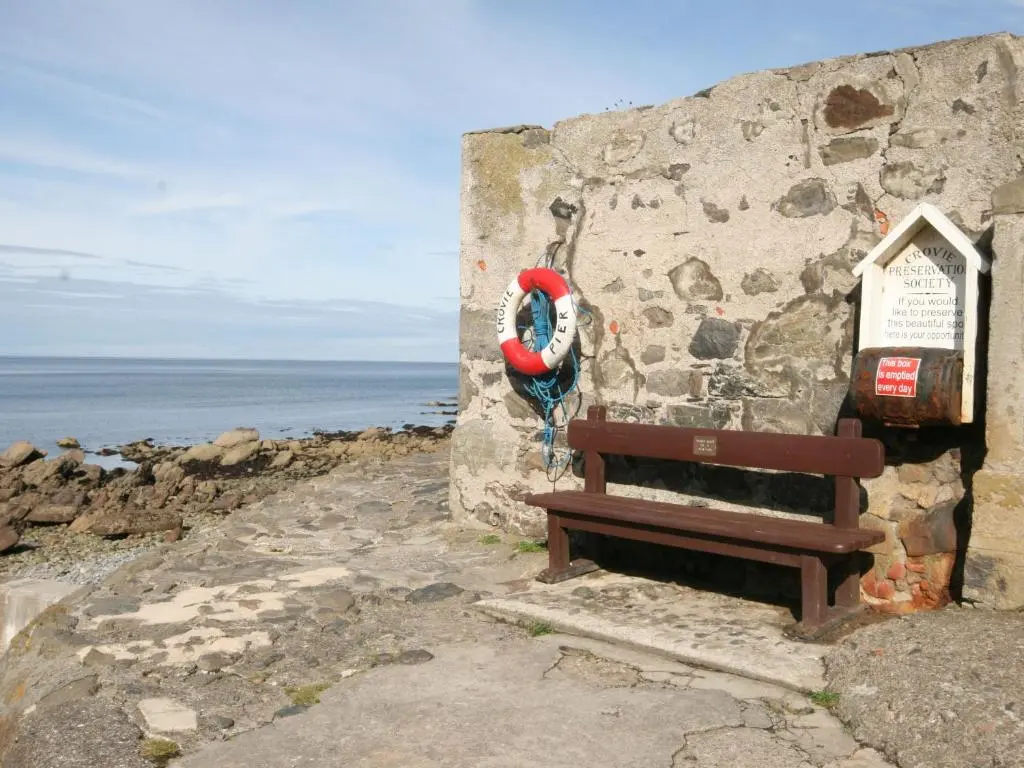 a bench sitting next to a stone wall next to the ocean at Crovie Cottage in Gardenstown
