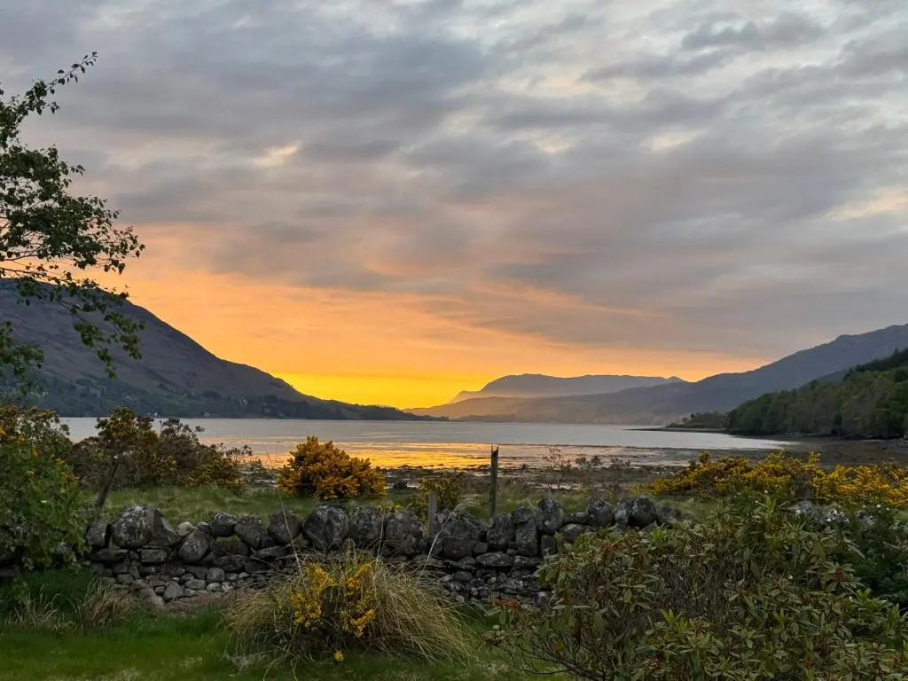 a sunset over a lake with a stone wall at Lochend Pods - Deas Pod in Inverlael