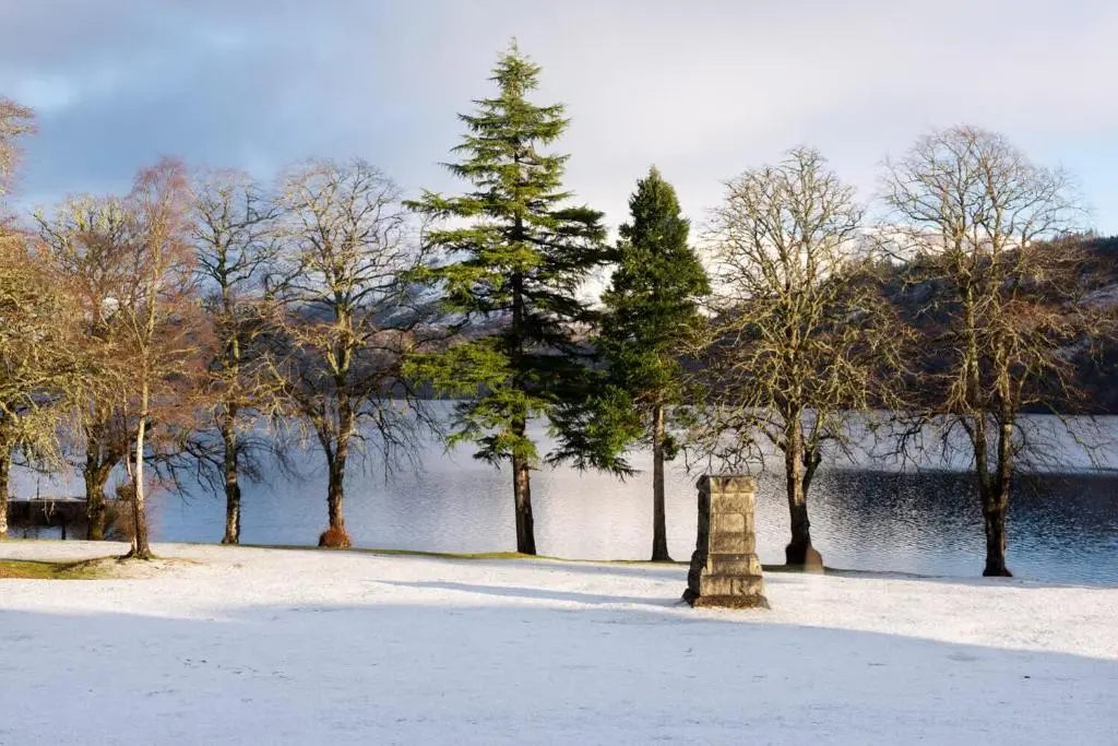 a snow covered park with trees and a body of water at Boys Dormitory in Fort Augustus