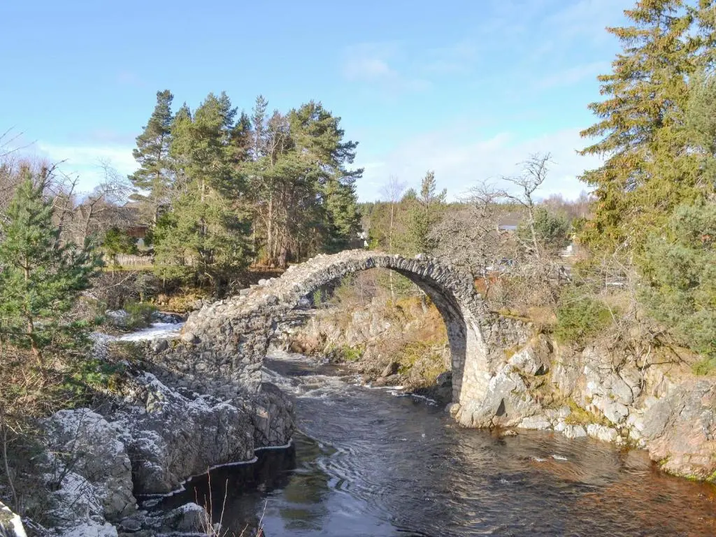 a stone bridge over a river in a forest at West End Cottage in Carrbridge