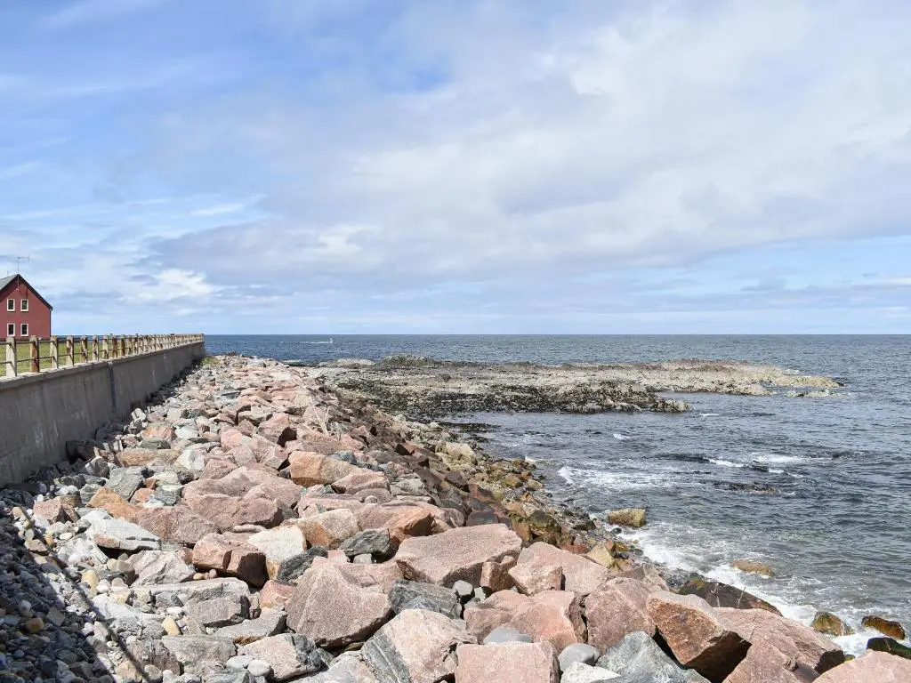 a stone wall next to the ocean with a red house at Sea Breeze Cottage in Buckie
